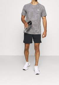 Grå camosmönstrad sportt-shirt, svarta shorts och vita sneakers. Håller en genomskinlig vattenflaska med svart lock.