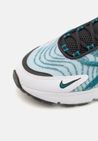 Gros plan sur la pointe d’une basket en cuir blanc, ornée de motifs vagues bleus, de lacets noirs en maille et d’un petit logo Nike virgule turquoise.