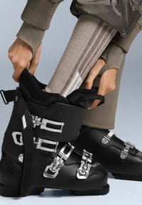 Svarta skidor med metallspännen och remmar, kombinerade med en texturerad beige strumpa med ljusa ränder, på en ljusgrå bakgrund.