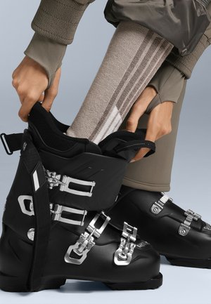 Crne skijaške čizme s metalnim kopčama i trakama, uparenim s teksturiranom bež čarapu s svijetlim prugama, na svijetlosivoj pozadini.