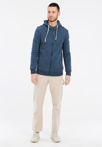 Námornícky zip-hoodie s kapucňou na šnúrku, texturovaná tkanina, rebrované manžety a spodný lem, spárované s béžovými nohavicami a bielymi teniskami.