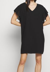 Svart klänning med V-ringning och korta, volangiga genomskinliga ärmar. Designen är löspassande med en mjuk textur och knälång fåll.