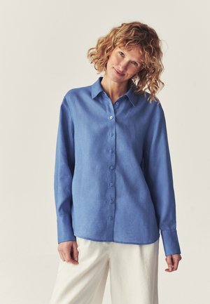 Woman with curly hair wearing a blue long-sleeve button-up shirt and white pants, standing against a plain light background.
