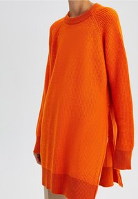 Luźny sweter z długim rękawem w żywym pomarańczowym kolorze, wykonany z dzianiny o fakturowanej powierzchni, z okrągłym dekoltem i bocznymi rozcięciami dla wygody.