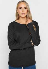 Schwarzes Langarmshirt mit einem Boot-Ausschnitt, glatter Textur, seitlichen Schlitzen und einer lockeren Passform, kombiniert mit blauen Jeans.