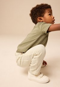 Zelena majica s kratkimi rokavi, bele črtaste hlače, beli supergi. Otrok v počepu na nevtralnem ozadju.