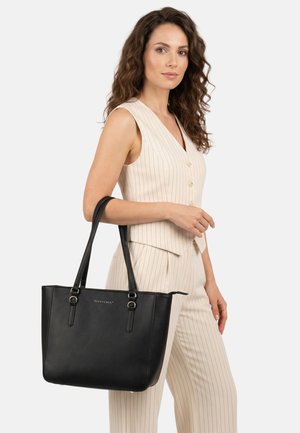 Frau in cremefarben gestreifter Weste und Hose, die eine große schwarze Ledertragetasche über der Schulter hält vor einem einfarbigen Hintergrund.