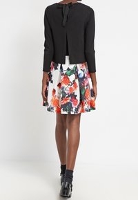 Svart kort jacka med en rosett baktill, kombinerad med en knälång blommig kjol i röd, grön och lila mönster. Svarta strumpor och skor.