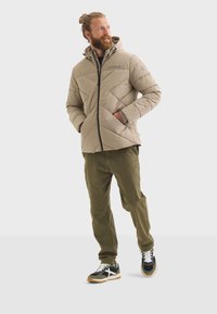 Chaqueta acolchada beige con capucha, cierre de cremallera, detalles en negro y bolsillos laterales. Combinada con pantalones verde oliva y zapatillas negras.