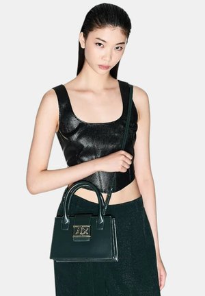 Top corto nero con texture abbinato a una borsa verde lucida con logo in metallo dorato. La borsa ha manici corti e forma rettangolare.