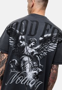 Czarny t-shirt z dużą grafiką anioła na plecach, z napisem "GOOD FOR NOTHING" w pogrubionych literach powyżej. Krótkie rękawy, okrągły dekolt.
