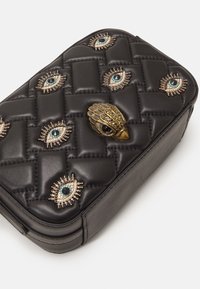 Clutch in pelle nera trapuntata decorato con motivi a occhio di gioiello e un fermaglio centrale a forma di testa d'uccello dorata su uno sfondo bianco.
