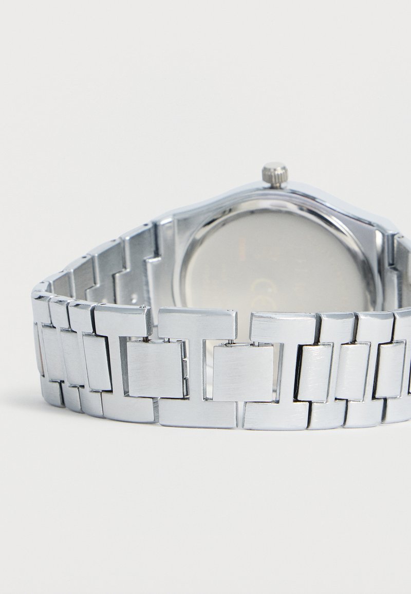Pulsera de reloj de eslabones de metal plateado con cierre, mostrada desde atrás con la esfera del reloj desenfocada.