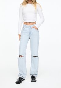 Vaqueros rasgados de un azul claro con un corte relajado y una cintura alta, combinados con una blusa ajustada de manga larga blanca y zapatos negros con tachuelas.