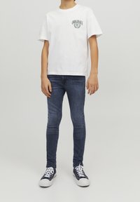 Camiseta blanca de algodón con texto verde "ORIGINALS", combinada con jeans ajustados azules y zapatillas marinas con suelas y cordones blancos.