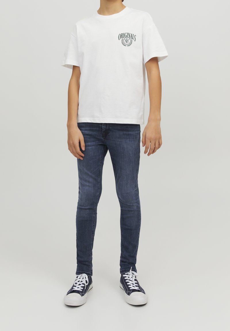Camiseta blanca de algodón con texto verde "ORIGINALS", combinada con jeans ajustados azules y zapatillas marinas con suelas y cordones blancos.