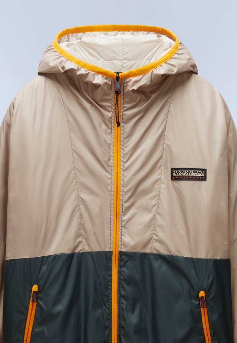Chaqueta ligera con la parte superior beige y la sección inferior en verde oscuro. Presenta un zipper naranja y un forro de capucha en el mismo color, además de bolsillos laterales.