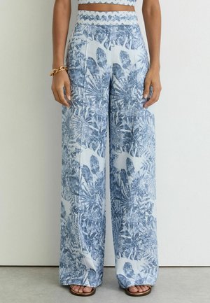 Vrouw draagt een broek met hoge taille en wijde pijpen met blauw-wit tropisch bladprint, gecombineerd met sandalen met open teen en een gouden armband.
