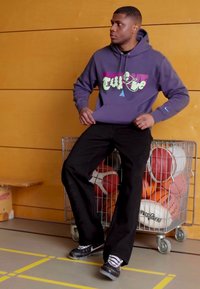 Felpa viola con design grafico multicolore, pantaloni neri larghi e sneaker nere. Sullo sfondo c'è un carrello di metallo con palle sportive.