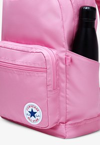 Roosa seljakott, millel on sile nailonist tekstuur, ees lukuga tasku ja küljetasku, kus hoitakse musta veepudelit. Kuvatud Converse'i logo.