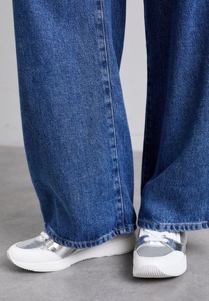 Sneakers sportivi argento e bianchi con dettagli in rete e lucidi, abbinati a jeans in denim blu a gamba larga con orlo grezzo.