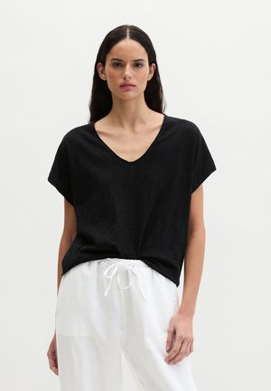 Woman with long dark hair wearing a loose black V-neck T-shirt and white drawstring pants, standing against a plain white background.