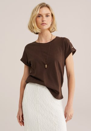 Blondínka v tmavohnedej krátkozásterovej košeli s gombíkmi a bielou textúrovanou sukňou, doplnená náhrdelníkom a prsteňmi.