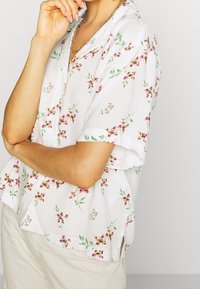 Personne avec les bras croisés portant une chemise blanche à manches courtes avec un motif floral rose et un pantalon beige, sur un fond neutre.