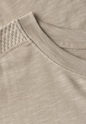 Detailní záběr béžové látky s texturovaným ramenním prostorem malými vystouplými vzory, hladkým výstřihem a viditelnými detaily šití.