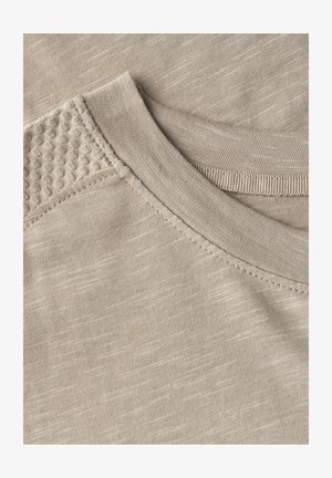 Zbliżenie beżowego materiału z fakturowanym ramieniem z małymi wypukłymi wzorami, gładką linią dekoltu i widocznymi detalami ściegów.