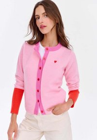 Różowy kardigan z kontrastującymi czerwonymi ściągaczami, z przodu zapinany na guziki, z małym czerwonym serduszkiem na klatce piersiowej. Miękka tekstura i dopasowany krój.