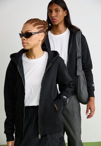 Sort zip-up hættetrøje båret over en hvid t-shirt, parret med grå bukser. Modellen har fletninger og solbriller, mens hun holder en ternet taske.