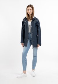 Marinblå vattentät jacka med huva, utrustad med silverfärgade tryckknappar och justerbar dragsko. Kombineras med ljusblå smala jeans och vita sneakers.