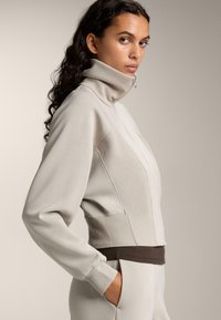 Svetlý sivý zipsový kabát s vysokým golierom, rebrovanou textúrou po bokoch a hnedým páskom. Má dlhé rukávy a bočné vrecká.