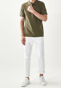 Polo shirt verde oliva con colletto e bordi bianchi, abbinato a jeans bianchi slim-fit e sneaker bianche, che mostrano un outfit casual.