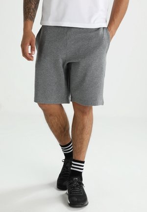 Mand iført grå knælange shorts, hvid skjorte, sorte sneakers og sorte sokker med hvide striber, stående med hænderne i lommerne.
