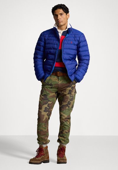 Casaco puffer azul com design matelassado, sobre um suéter listrado em vermelho e azul-marinho. Calças cargo em padrão camuflado e botas de caminhada castanhas completam o visual.
