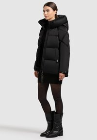 Naine kannab musta paksu jope suurt kraega, musta kudumist kleiti, läbipaistvaid sukkpükse ja musti kuni pahkluuni saapaid, seistes külg ees halli tausta taustal.