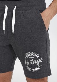 Shorts en coton gris foncé avec une taille élastique et des cordons blancs. Présente un imprimé graphique de style vintage en blanc sur le côté.