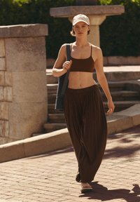Jeune femme marchant à l'extérieur, portant un crop top marron, un pantalon marron ample, une casquette blanche, des sandales et un sac noir pour tapis de yoga.