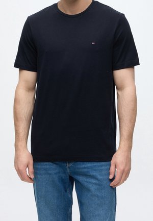 Homme portant un t-shirt noir uni à manches courtes et un jean bleu, debout devant un fond blanc uni.