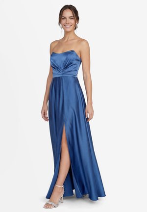 Lächelnde Frau in einem trägerlosen, blauen Satinkleid mit Schlitz vorne und silbernen High-Heel-Sandalen, stehend vor weißem Hintergrund.