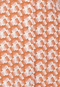 Camisa de botões com um padrão floral laranja e branco, intercalado com formas geométricas, feita de tecido leve, com botões brancos.