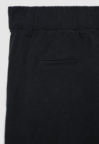 Schwarze gerippte Hose mit Gürtelschlaufen und einer einzigen Paspeltasche auf der Rückseite, die die detaillierte Stoffstruktur zeigt.