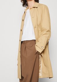 Persoon in een beige trenchcoat, witte t-shirt en bruine geplooide broek met een hand in de zak, tegen een effen achtergrond.