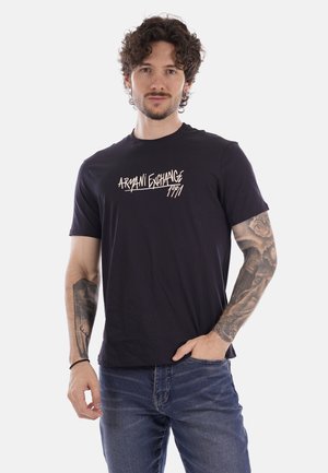 Hombre con cabello rizado y tatuajes en ambos brazos lleva una camiseta negra de Armani Exchange y vaqueros azules, posando frente a un fondo blanco.