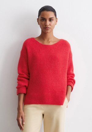 Red knit sweater with a wide neckline and ribbed texture. Slouchy fit with rolled sleeves. Paired with light-colored trousers.