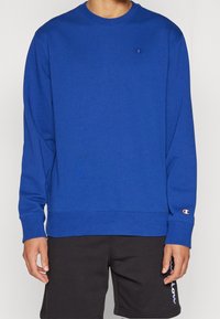 Blå sweatshirt med långa ärmar och rund halsringning. Har en logotyp på bröstet samt ribbade muddarna och nederkanten. Matchas med svarta shorts.