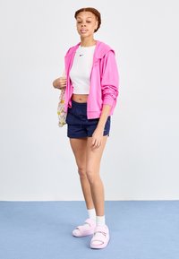 Felpa rosa con zip sopra un top bianco corto, pantaloni corti blu navy e sandali rosa con accenti bianchi. Outfit atletico semplice e casual.