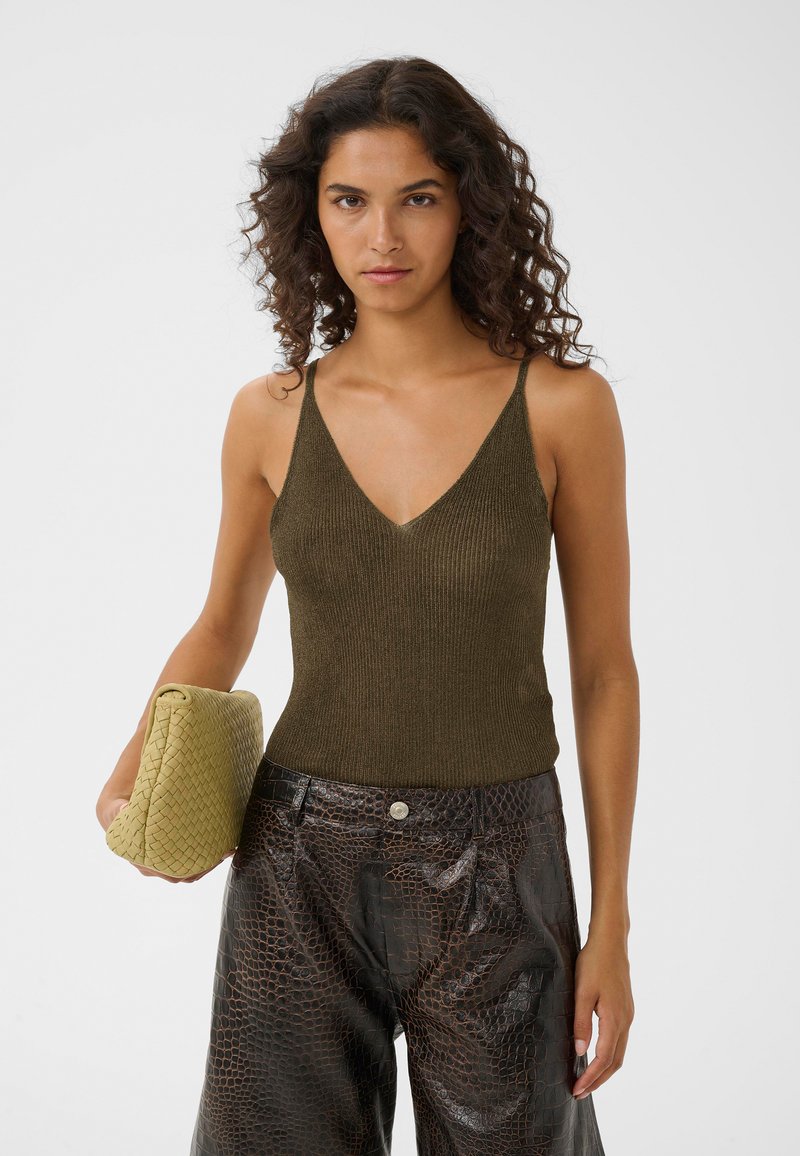 Γυναίκα με σγουρά μαλλιά, που φοράει πράσινο ελαιώδες πλεκτό τοπ και σκούρα καφέ παντελόνια από συνθετικό δέρμα κροκόδειλου, κρατώντας ένα μεγάλο πλεκτό μπεζ τσαντάκι.